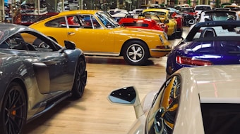 A collection of luxury sports cars are displayed in an indoor showroom with a polished wooden floor. The focus is on a vintage yellow car surrounded by modern vehicles of various colors, including red, blue, and gray. The setting appears affluent and neatly organized, highlighting the elegance and design of each car.