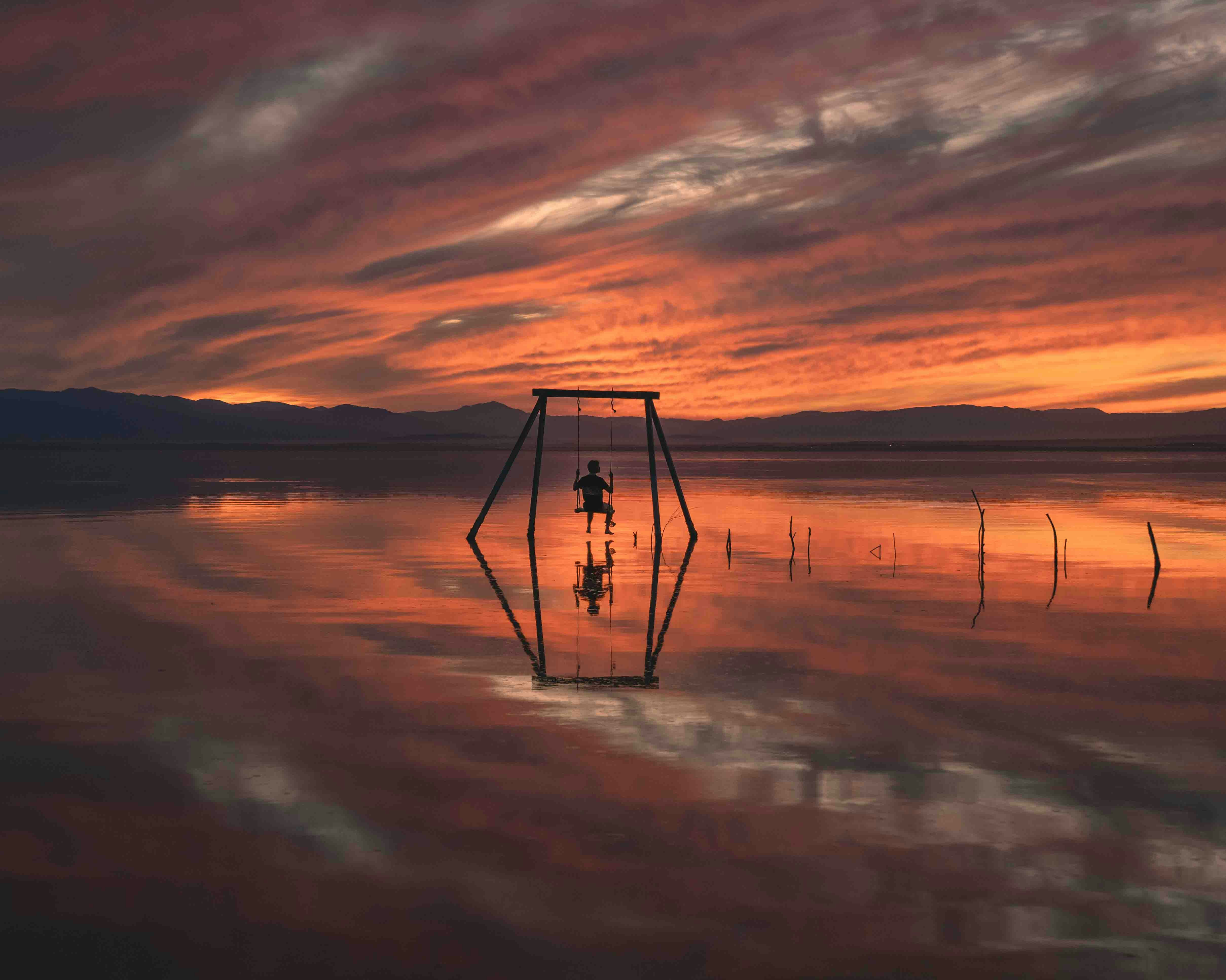 On a swing set up in a water body, we see the silhouette of a person swinging. Mountains line the water, and the sky is thick with clouds, golden in twilight hours. 