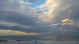 A serene ocean scene features a wide expanse of water with gentle waves under a vast sky filled with a blend of dark and light clouds, highlighting various shades of blue and grey. Soft sunlight breaks through, casting a subtle glow onto the clouds and water.
