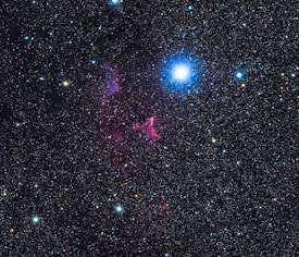 A vast starry sky filled with numerous stars, with a particularly prominent bright blue star near the center. Wisps of pink and purple nebulous clouds are scattered amidst the stars, adding a sense of depth and cosmic wonder.