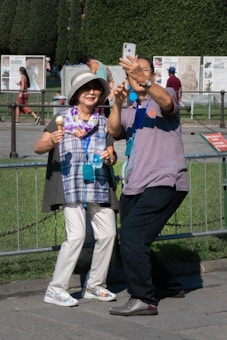 A couple is taking a selfie outdoors in a sunny setting. The woman is wearing a large sunhat, colorful scarf, and is holding an ice cream cone and a water bottle. The man is holding a smartphone, capturing the moment. They are in a public area with grass, informational boards, and other people in the background.