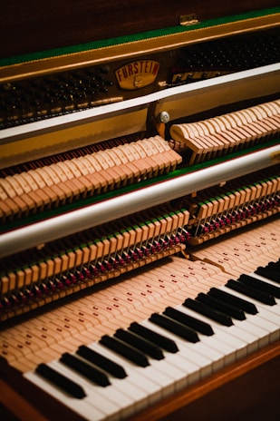 The image displays the interior mechanism of an upright piano, focusing on the hammers, strings, and the keyboard. The wooden hammers are aligned in rows, and the black and white keys of the keyboard are visible at the bottom.