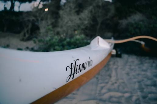 A close-up of a white canoe or kayak with the word 'Honu Vela' inscribed on its side. The background features a sandy beach and thick, green foliage, slightly out of focus, suggesting a natural setting.