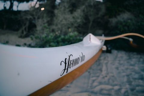 A close-up of a white canoe or kayak with the word 'Honu Vela' inscribed on its side. The background features a sandy beach and thick, green foliage, slightly out of focus, suggesting a natural setting.