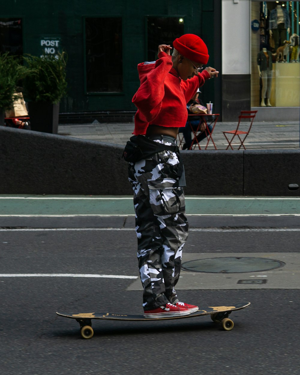 uomo in felpa con cappuccio rossa e pantaloni mimetici in bianco e nero in piedi sullo skateboard rosso durante il giorno