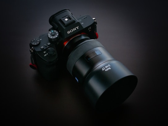 A high-quality digital camera with a large lens is placed against a dark background. The camera body features several control dials and buttons, along with clear branding. The lens appears to be a professional-grade, emphasizing optical precision.