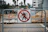 A barrier sign with a 'no pedestrian' symbol is positioned in front of a construction site. Behind the sign, there are construction materials, orange cones, and a chain-link fence. In the background, palm trees and buildings are visible.