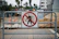 A barrier sign with a 'no pedestrian' symbol is positioned in front of a construction site. Behind the sign, there are construction materials, orange cones, and a chain-link fence. In the background, palm trees and buildings are visible.