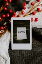A close-up of a Polaroid photo with the word 'memories' written on it, placed on a textured dark fabric with festive red berry branches. Soft, knitted white fabric partially drapes the scene, adding contrast and warmth.