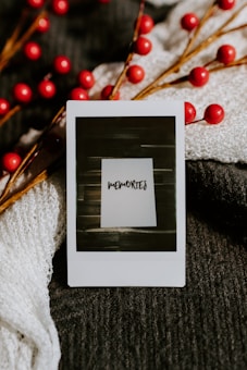 A close-up of a Polaroid photo with the word 'memories' written on it, placed on a textured dark fabric with festive red berry branches. Soft, knitted white fabric partially drapes the scene, adding contrast and warmth.