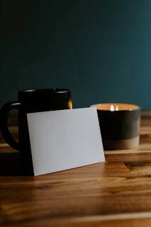 A wooden surface holds a dark ceramic mug, a lit candle with a visible flame, and a blank white card propped upright. The background is a dark muted color, enhancing the warm and cozy atmosphere.