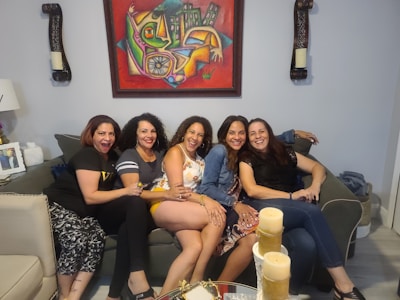 Five women are sitting closely together on a couch, smiling and laughing. They appear to be enjoying each other's company. Behind them is a colorful abstract painting. There are decorative items on the walls and a lamp on the left side.
