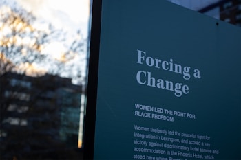 A sign with text in white on a dark background that reads 'Forcing a Change' and details the role of women in leading the fight for black freedom. The background includes a blurred outdoor scene with trees and a building.