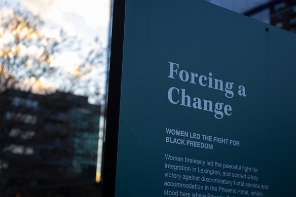 A sign with text in white on a dark background that reads 'Forcing a Change' and details the role of women in leading the fight for black freedom. The background includes a blurred outdoor scene with trees and a building.