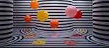 The image features several vibrant, colorful balloons floating against a backdrop of black and white striped walls and floors. The stripes follow a wavy pattern that extends onto the floor, creating a visually striking and playful effect. The color palette of the balloons includes orange, yellow, red, and pink, adding a pop of color to the monochromatic background.
