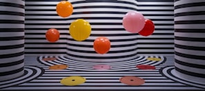 The image features several vibrant, colorful balloons floating against a backdrop of black and white striped walls and floors. The stripes follow a wavy pattern that extends onto the floor, creating a visually striking and playful effect. The color palette of the balloons includes orange, yellow, red, and pink, adding a pop of color to the monochromatic background.