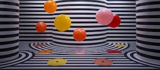 The image features several vibrant, colorful balloons floating against a backdrop of black and white striped walls and floors. The stripes follow a wavy pattern that extends onto the floor, creating a visually striking and playful effect. The color palette of the balloons includes orange, yellow, red, and pink, adding a pop of color to the monochromatic background.