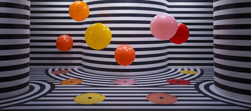 The image features several vibrant, colorful balloons floating against a backdrop of black and white striped walls and floors. The stripes follow a wavy pattern that extends onto the floor, creating a visually striking and playful effect. The color palette of the balloons includes orange, yellow, red, and pink, adding a pop of color to the monochromatic background.