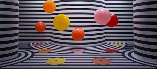 The image features several vibrant, colorful balloons floating against a backdrop of black and white striped walls and floors. The stripes follow a wavy pattern that extends onto the floor, creating a visually striking and playful effect. The color palette of the balloons includes orange, yellow, red, and pink, adding a pop of color to the monochromatic background.