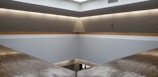 A minimalist interior space with a high ceiling and light gray walls. A horizontal wooden beam divides the upper and lower sections. Recessed lighting around the ceiling creates a soft, warm glow. The architecture features clean lines and a modern design with an open and spacious feel.