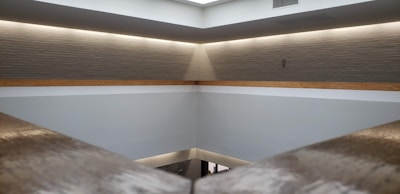 A minimalist interior space with a high ceiling and light gray walls. A horizontal wooden beam divides the upper and lower sections. Recessed lighting around the ceiling creates a soft, warm glow. The architecture features clean lines and a modern design with an open and spacious feel.