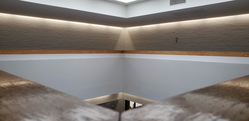 A minimalist interior space with a high ceiling and light gray walls. A horizontal wooden beam divides the upper and lower sections. Recessed lighting around the ceiling creates a soft, warm glow. The architecture features clean lines and a modern design with an open and spacious feel.