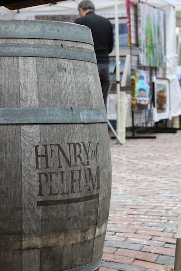A large wooden barrel inscribed with 'Henry of Pelham' stands prominently in the foreground on a cobblestone surface. In the background, a person is slightly blurred, standing beside a stall or display of artwork or photographs.