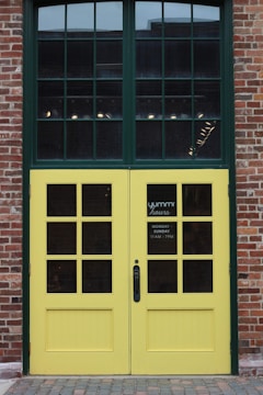 A double door with a grid window design, painted yellow and framed by red brick walls. The top section consists of large window panes, while the lower section has smaller square windows. A sign on one door indicates the business hours from Monday to Sunday, 11 AM to 7 PM.