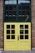 A double door with a grid window design, painted yellow and framed by red brick walls. The top section consists of large window panes, while the lower section has smaller square windows. A sign on one door indicates the business hours from Monday to Sunday, 11 AM to 7 PM.