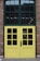 A double door with a grid window design, painted yellow and framed by red brick walls. The top section consists of large window panes, while the lower section has smaller square windows. A sign on one door indicates the business hours from Monday to Sunday, 11 AM to 7 PM.