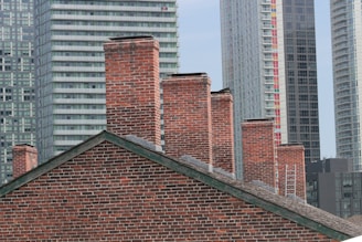 A collection of red brick chimneys on a slanted rooftop is set against the background of modern high-rise buildings. The contrast between the old-style brick architecture and the contemporary glass and steel skyscrapers creates a striking juxtaposition.