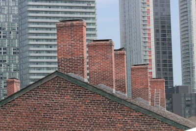 A collection of red brick chimneys on a slanted rooftop is set against the background of modern high-rise buildings. The contrast between the old-style brick architecture and the contemporary glass and steel skyscrapers creates a striking juxtaposition.