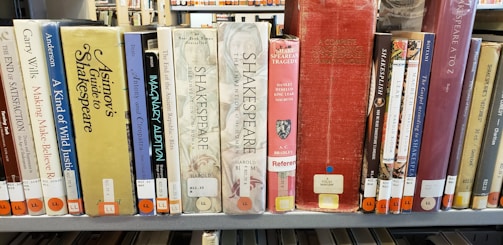 A row of books on a library shelf, featuring various titles related to Shakespeare and other literary topics. The books are in a mix of colors, and there are labels on the spines indicating their library classification.