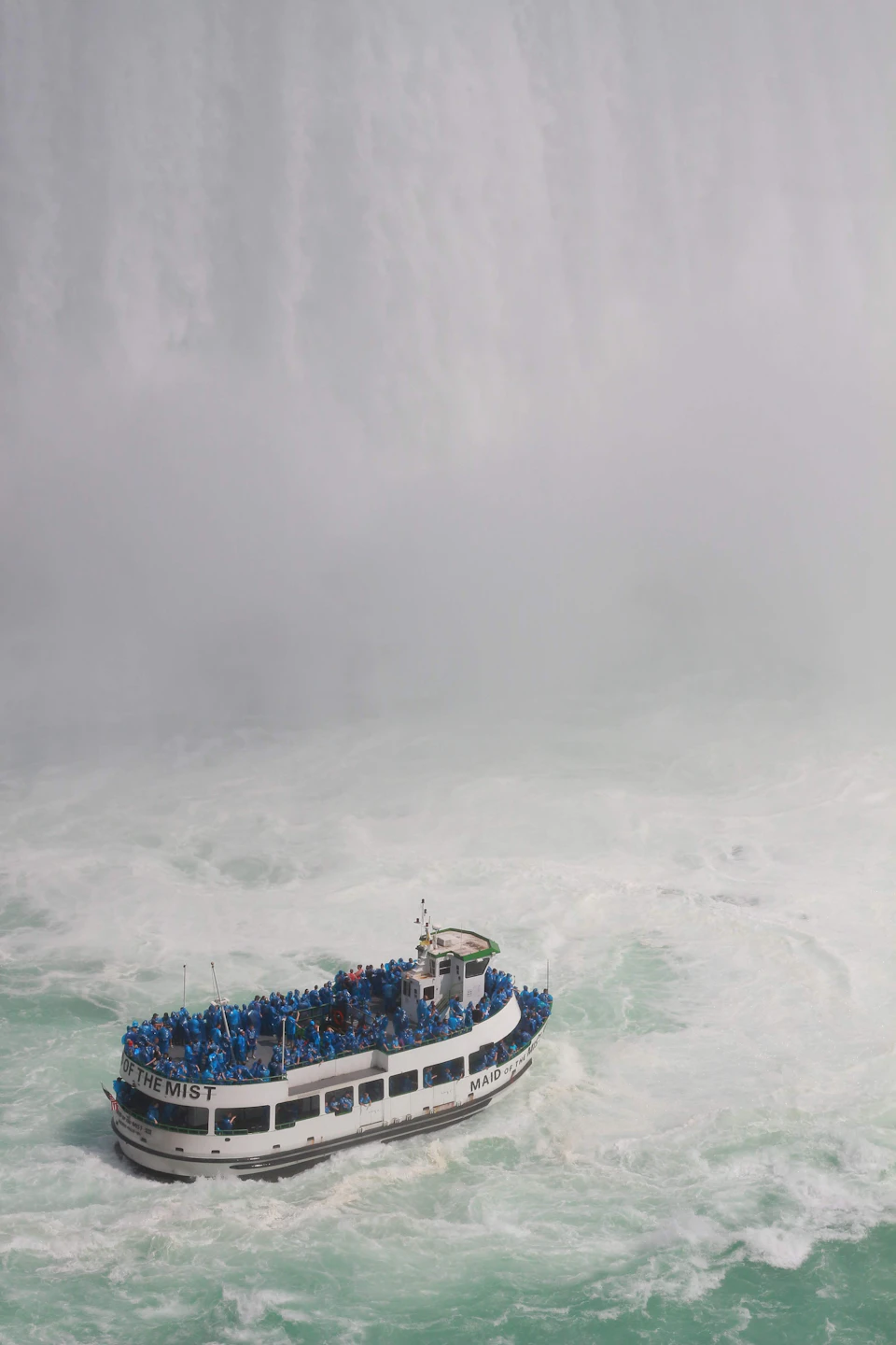 The image depicts a large white boat with blue accents, filled with people enjoying their time on board. The boat is surrounded by choppy green waters and appears to be moving towards the right side of the frame. In the background, a waterfall can be seen, adding to the natural beauty of the scene.