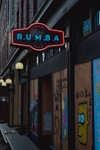 A dimly lit street scene features a neon sign displaying the word 'RUMBA' in blue lettering with a red outline. The surroundings are urban, with brick walls and boarded-up windows adorned with colorful graffiti, including a character with large eyes. Light fixtures hang along the building's facade.