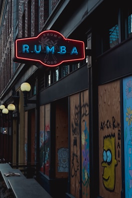 A dimly lit street scene features a neon sign displaying the word 'RUMBA' in blue lettering with a red outline. The surroundings are urban, with brick walls and boarded-up windows adorned with colorful graffiti, including a character with large eyes. Light fixtures hang along the building's facade.