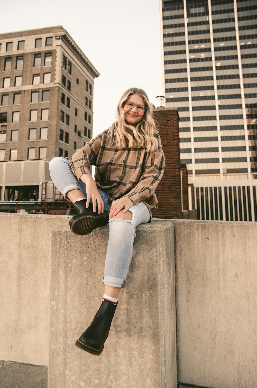 woman in brown leather jacket and blue denim jeans sitting on concrete wall during daytime