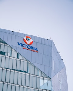 Modern multi-story building with a prominent sign for Victoria Medical Center on its upper facade. The building features sleek glass and metal architecture with evenly spaced windows, and the sky in the background is clear and blue.