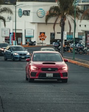 A red sports car drives on a city street, followed by other vehicles. Palm trees are visible along the road, with a commercial building featuring the AT&T logo in the background. Motorcycles are parked nearby, and the general scene suggests a busy urban environment.