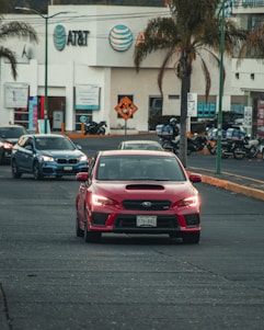 A red sports car drives on a city street, followed by other vehicles. Palm trees are visible along the road, with a commercial building featuring the AT&T logo in the background. Motorcycles are parked nearby, and the general scene suggests a busy urban environment.