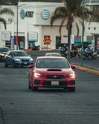 A red sports car drives on a city street, followed by other vehicles. Palm trees are visible along the road, with a commercial building featuring the AT&T logo in the background. Motorcycles are parked nearby, and the general scene suggests a busy urban environment.