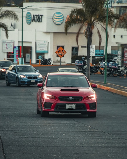 A red sports car drives on a city street, followed by other vehicles. Palm trees are visible along the road, with a commercial building featuring the AT&T logo in the background. Motorcycles are parked nearby, and the general scene suggests a busy urban environment.