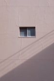 A simple beige wall with a small rectangular window, featuring a sliding design. The wall's texture appears rough and paint has subtle cracks. A shadow, likely from a utility line or similar object, diagonally crosses the bottom section of the image.
