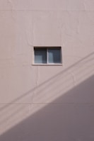A simple beige wall with a small rectangular window, featuring a sliding design. The wall's texture appears rough and paint has subtle cracks. A shadow, likely from a utility line or similar object, diagonally crosses the bottom section of the image.