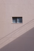 A simple beige wall with a small rectangular window, featuring a sliding design. The wall's texture appears rough and paint has subtle cracks. A shadow, likely from a utility line or similar object, diagonally crosses the bottom section of the image.