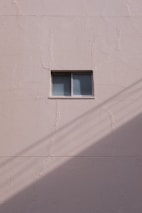 A simple beige wall with a small rectangular window, featuring a sliding design. The wall's texture appears rough and paint has subtle cracks. A shadow, likely from a utility line or similar object, diagonally crosses the bottom section of the image.