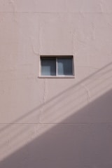 A simple beige wall with a small rectangular window, featuring a sliding design. The wall's texture appears rough and paint has subtle cracks. A shadow, likely from a utility line or similar object, diagonally crosses the bottom section of the image.