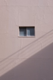 A simple beige wall with a small rectangular window, featuring a sliding design. The wall's texture appears rough and paint has subtle cracks. A shadow, likely from a utility line or similar object, diagonally crosses the bottom section of the image.