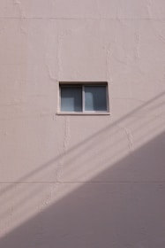 A simple beige wall with a small rectangular window, featuring a sliding design. The wall's texture appears rough and paint has subtle cracks. A shadow, likely from a utility line or similar object, diagonally crosses the bottom section of the image.