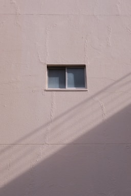 A simple beige wall with a small rectangular window, featuring a sliding design. The wall's texture appears rough and paint has subtle cracks. A shadow, likely from a utility line or similar object, diagonally crosses the bottom section of the image.